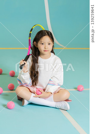 A young girl sits on a blue sports court with tennis balls around her. She holds a racket in one hand and a ball in the other. She looks serious and ready to play tennis. 139259641