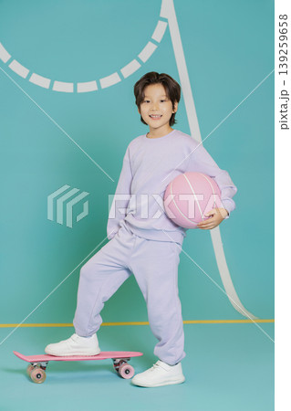 A child poses on a skateboard while holding a pink basketball in a brightly colored studio. The background features a simple design that adds to the fun atmosphere. A child poses on a skateboard while holding a pink basketball in a brightly colored studio. The background features a simple design that adds to the fun atmosphere. 139259658
