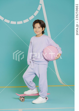 A child stands on a skateboard while holding a pink basketball in a brightly colored indoor space. The background shows a sports court with lines. It is a fun time for play. A child stands on a skateboard while holding a pink basketball in a brightly colored indoor space. The background shows a sports court with lines. It is a fun time for play. 139259661