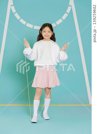A girl stands on a gym floor holding a skipping rope. She wears a white sweatshirt and a pink skirt. The area has blue walls with markings and shows a fun activity for children. A girl stands on a gym floor holding a skipping rope. She wears a white sweatshirt and a pink skirt. The area has blue walls with markings and shows a fun activity for children. 139259682
