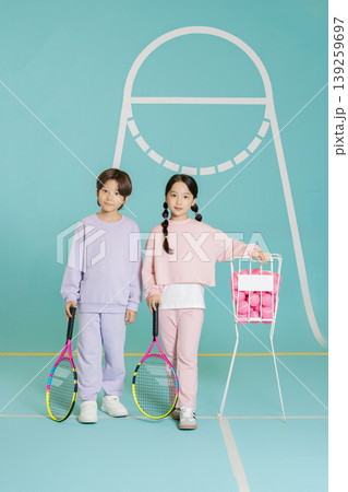 Two kids are on a tennis court. One child holds a racket and a basket of tennis balls. The other child also has a racket in hand. They look ready for practice. 139259697