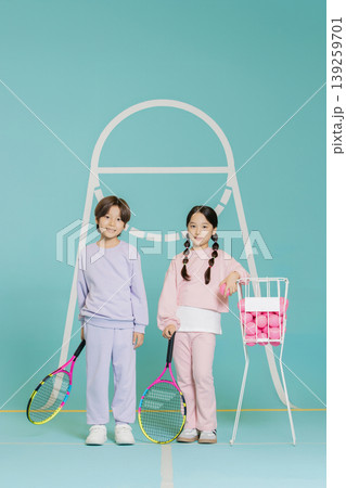 Two children stand on a tennis court with rackets in hand. One child holds a basket of tennis balls. They are smiling and ready to play tennis in a bright and colorful space. 139259701