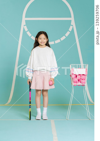 A young girl stands on a tennis court with a racket in one hand and a tennis ball in the other. A basket filled with pink tennis balls is next to her. She appears ready to play. 139259706