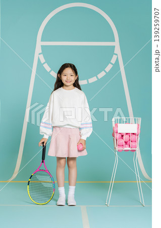 A girl holds a tennis racket in one hand and a tennis ball in the other. She stands next to a basket filled with bright tennis balls. The background shows a simple court design. A girl holds a tennis racket in one hand and a tennis ball in the other. She stands next to a basket filled with bright tennis balls. The background shows a simple court design. 139259707