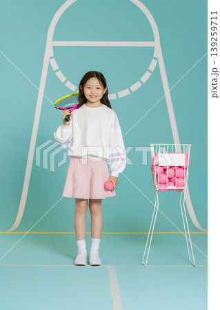 A girl stands in a tennis court setting. She holds a racket in one hand and a pink ball in the other. Behind her is a container filled with more pink balls. A girl stands in a tennis court setting. She holds a racket in one hand and a pink ball in the other. Behind her is a container filled with more pink balls. 139259711