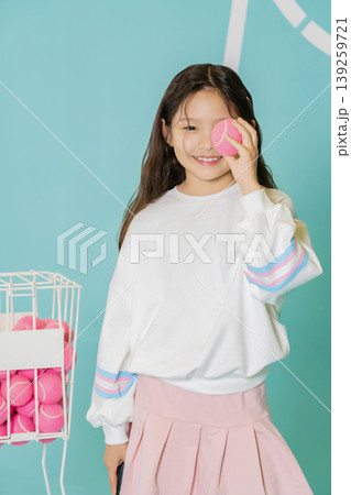 A child holds a pink tennis ball to her eye and smiles. She stands beside a wire basket filled with more pink balls. The background shows bright colors, creating a fun atmosphere. A child holds a pink tennis ball to her eye and smiles. She stands beside a wire basket filled with more pink balls. The background shows bright colors, creating a fun atmosphere. 139259721