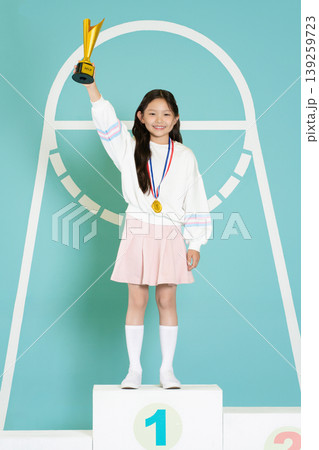 A young girl stands on a podium holding a trophy high with a big smile. She wears a medal around her neck and a sports outfit. The background shows a simple design for an event. 139259723