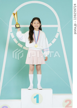 A girl stands on the first place podium holding a trophy with a medal around her neck. She smiles proudly wearing a simple outfit, showing her achievement in a competition. 139259724