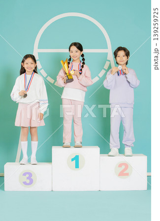 Three children stand on a podium, smiling and holding medals and trophies. They display their awards in a competition setting. The background is bright and colorful, adding to the joyful atmosphere. Three children stand on a podium, smiling and holding medals and trophies. They display their awards in a competition setting. The background is bright and colorful, adding to the joyful atmosphere. 139259725