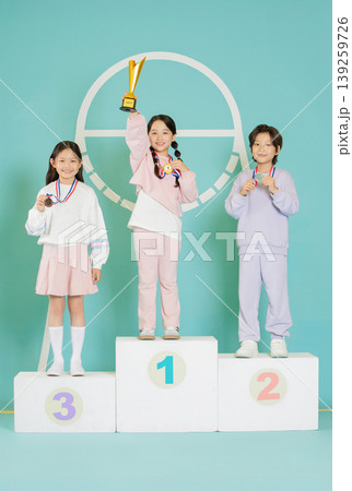 Three kids stand on a podium celebrating their success. One child holds a trophy high, while others show their medals. The backdrop is colorful and cheerful. 139259726