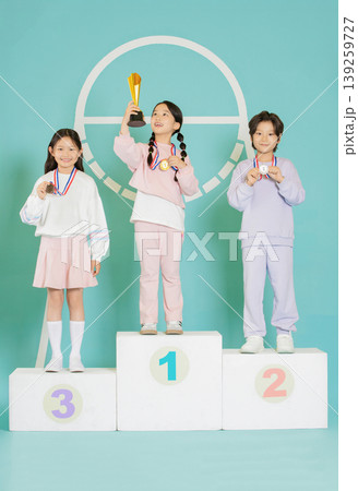 Three children celebrate their achievements on a podium. One child holds a trophy, while the others display their medals. They are in a colorful indoor setting. 139259727