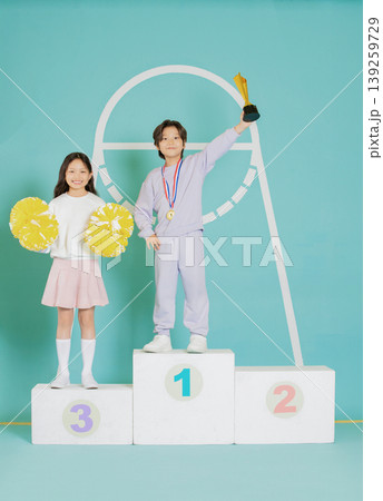 Two children stand on a podium after winning a competition. One child holds a trophy, while the other displays pom poms. The setting is bright and colorful, highlighting their achievements. Two children stand on a podium after winning a competition. One child holds a trophy, while the other displays pom poms. The setting is bright and colorful, highlighting their achievements. 139259729