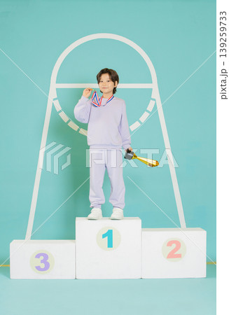 A young athlete stands proudly on a podium after winning a competition. They hold a trophy in one hand and touch their medal with the other. The background has a large hoop design. A young athlete stands proudly on a podium after winning a competition. They hold a trophy in one hand and touch their medal with the other. The background has a large hoop design. 139259733