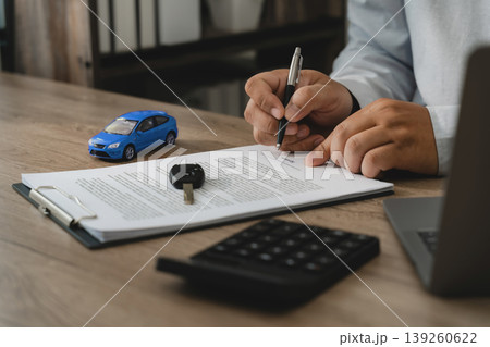 Man signing car insurance policy document with pen beside car key, model vehicle, calculator and laptop on desk, representing policy approval, coverage agreement, financial paperwork and auto contract 139260622