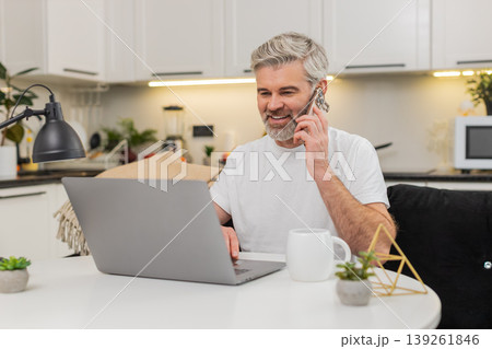 Middle-aged man on laptop handles call on smartphone discusses project tasks and writes updates live 139261846