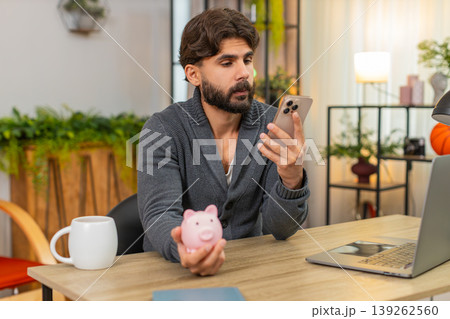 Indian man opens empty piggy bank sighs writes monthly budget plan in smartphone saving money goals Indian man opens empty piggy bank sighs writes monthly budget plan in smartphone saving money goals 139262560