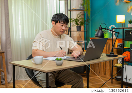Asian man taking notes from online course on laptop, explaining to camera with clear confident tone Asian man taking notes from online course on laptop, explaining to camera with clear confident tone 139262611
