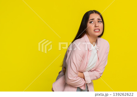 Young woman bending forward rubbing stomach showing painful expression suffering from abdominal ache Young woman bending forward rubbing stomach showing painful expression suffering from abdominal ache 139262622