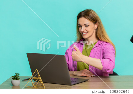 Adult woman sitting at table showing thumbs up to laptop webcam giving positive feedback with smile 139262643