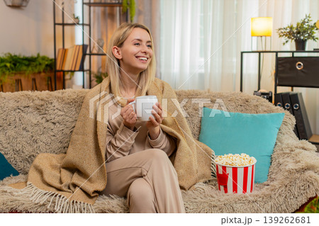 Adult woman wrapped in blanket sitting on sofa with cup of tea watching series thoughtfully at home Adult woman wrapped in blanket sitting on sofa with cup of tea watching series thoughtfully at home 139262681