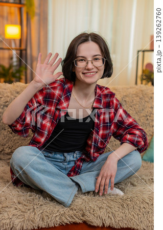 Young woman smiling and waving hand at home, greeting someone with friendly and warm expression Young woman smiling and waving hand at home, greeting someone with friendly and warm expression 139262760