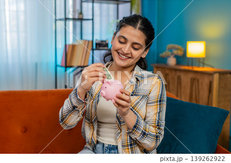 Indian woman thoughtfully count cash dollar money placing bills in piggy bank saving limited budget Indian woman thoughtfully count cash dollar money placing bills in piggy bank saving limited budget 139262922