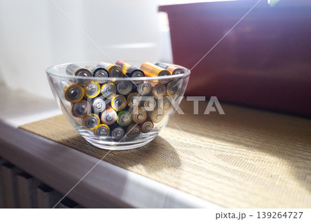 A young woman places a glass bowl filled with used batteries onto a wooden shelf for future disposal. This process demonstrates conscious household waste sorting and care for the environment. A young woman places a glass bowl filled with used batteries onto a wooden shelf for future disposal. This process demonstrates conscious household waste sorting and care for the environment. 139264727