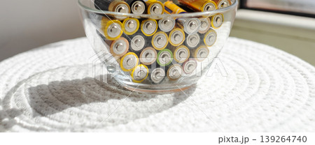 A young woman places a glass bowl filled with used batteries onto a wooden shelf for future disposal. This process demonstrates conscious household waste sorting and care for the environment. A young woman places a glass bowl filled with used batteries onto a wooden shelf for future disposal. This process demonstrates conscious household waste sorting and care for the environment. 139264740