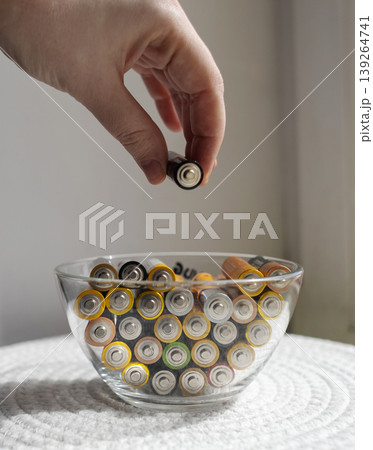 A young woman places a glass bowl filled with used batteries onto a wooden shelf for future disposal. This process demonstrates conscious household waste sorting and care for the environment. A young woman places a glass bowl filled with used batteries onto a wooden shelf for future disposal. This process demonstrates conscious household waste sorting and care for the environment. 139264741
