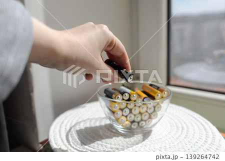 A young woman places a glass bowl filled with used batteries onto a wooden shelf for future disposal. This process demonstrates conscious household waste sorting and care for the environment. A young woman places a glass bowl filled with used batteries onto a wooden shelf for future disposal. This process demonstrates conscious household waste sorting and care for the environment. 139264742