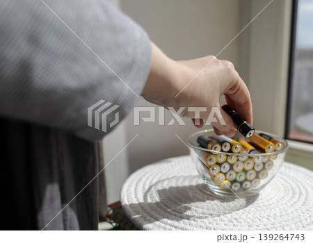 A young woman places a glass bowl filled with used batteries onto a wooden shelf for future disposal. This process demonstrates conscious household waste sorting and care for the environment. 139264743