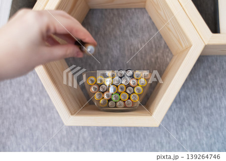 A young woman places a glass bowl filled with used batteries onto a wooden shelf for future disposal. This process demonstrates conscious household waste sorting and care for the environment. A young woman places a glass bowl filled with used batteries onto a wooden shelf for future disposal. This process demonstrates conscious household waste sorting and care for the environment. 139264746