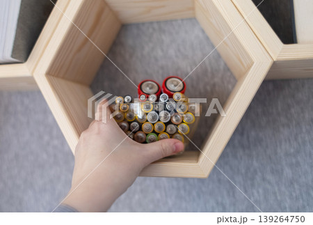 A young woman places a glass bowl filled with used batteries onto a wooden shelf for future disposal. This process demonstrates conscious household waste sorting and care for the environment. A young woman places a glass bowl filled with used batteries onto a wooden shelf for future disposal. This process demonstrates conscious household waste sorting and care for the environment. 139264750