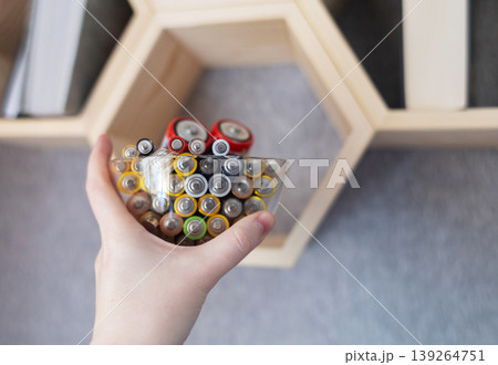 A young woman places a glass bowl filled with used batteries onto a wooden shelf for future disposal. This process demonstrates conscious household waste sorting and care for the environment. A young woman places a glass bowl filled with used batteries onto a wooden shelf for future disposal. This process demonstrates conscious household waste sorting and care for the environment. 139264751