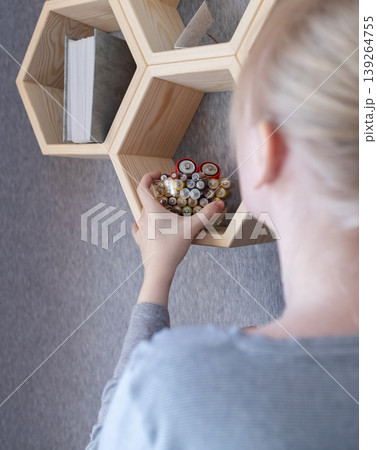 A young woman places a glass bowl filled with used batteries onto a wooden shelf for future disposal. This process demonstrates conscious household waste sorting and care for the environment. A young woman places a glass bowl filled with used batteries onto a wooden shelf for future disposal. This process demonstrates conscious household waste sorting and care for the environment. 139264755