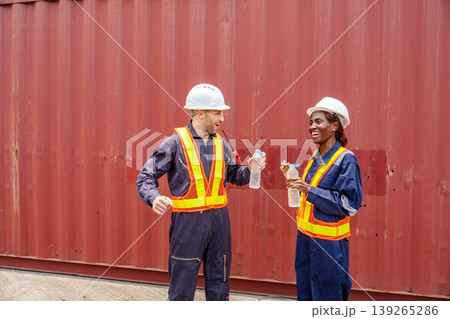 Smiling mid-adult Caucasian male and Black female industrial workers sharing a laugh while drinking water at an Asian container yard. 139265286