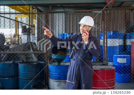 Mid-adult Caucasian male logistics coordinator in navy coveralls pointing and using a walkie-talkie at an Asian industrial warehouse. Mid-adult Caucasian male logistics coordinator in navy coveralls pointing and using a walkie-talkie at an Asian industrial warehouse. 139265290