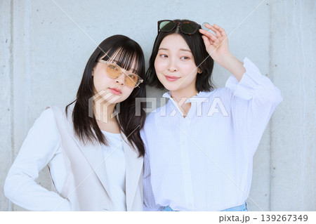 サングラスをかけた白い服の若い女性二人がコンクリート壁の前でポーズをとる友達同士のポートレート サングラスをかけた白い服の若い女性二人がコンクリート壁の前でポーズをとる友達同士のポートレート 139267349