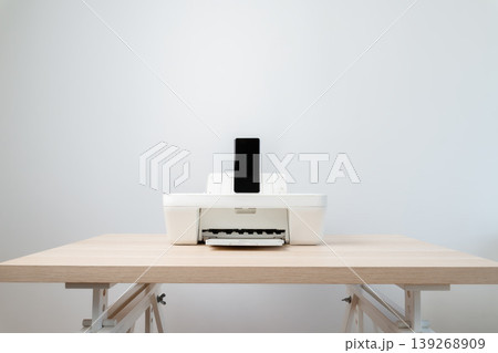 Minimal white printer with a smartphone docked on a light wooden desk, perfect for modern home office, wireless printing, technology convenience and clean workspace concept photography. Minimal white printer with a smartphone docked on a light wooden desk, perfect for modern home office, wireless printing, technology convenience and clean workspace concept photography. 139268909