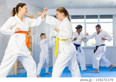 Young girl and woman in kimono and colored belt practicing karate punch block during group martial arts lesson in gym Young girl and woman in kimono and colored belt practicing karate punch block during group martial arts lesson in gym 139272179
