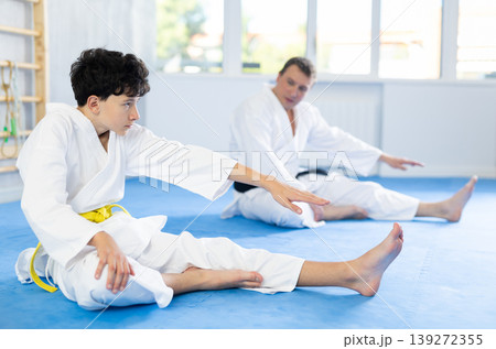 Teenage boy and middle-aged man trainer wearing kimono doing leg stretching in sports hall 139272355