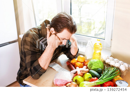 Stressed man with empty wallet and long receipt sits by groceries. Concept of high food prices, inflation, and economic crisis impact on household budget. Stressed man with empty wallet and long receipt sits by groceries. Concept of high food prices, inflation, and economic crisis impact on household budget. 139272454