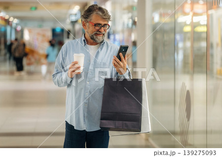 Cup of drink and smartphone are in hands. Senior man is in the mall, shopping time conception Cup of drink and smartphone are in hands. Senior man is in the mall, shopping time conception 139275093