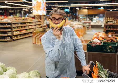Making fun face with banana. A senior man is buying food in the grocery store Making fun face with banana. A senior man is buying food in the grocery store 139275239