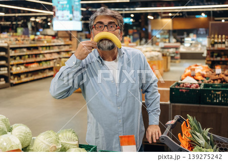 Making fun face with banana. A senior man is buying food in the grocery store Making fun face with banana. A senior man is buying food in the grocery store 139275242