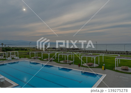 Calm evening by the seaside pool under a cloudy sky with distant mountains Calm evening by the seaside pool under a cloudy sky with distant mountains 139275278