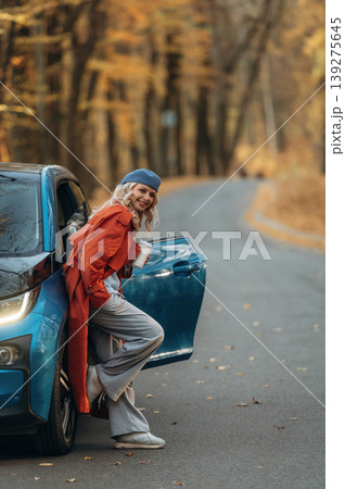 Smiling, having fun. Beautiful blonde woman is with her blue electric car on the road of forest 139275645