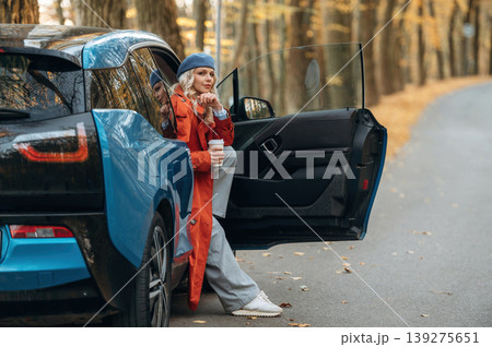 Door is opened, sitting and holding drink. Beautiful blonde woman is with her blue electric car on the road of forest 139275651