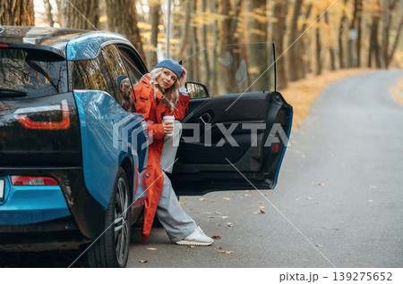 Door is opened, sitting and holding drink. Beautiful blonde woman is with her blue electric car on the road of forest 139275652