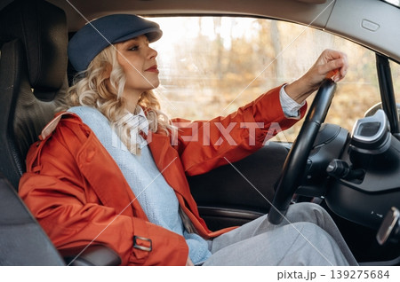 In red coat and blue beret. Happy blonde woman is sitting her electric car In red coat and blue beret. Happy blonde woman is sitting her electric car 139275684